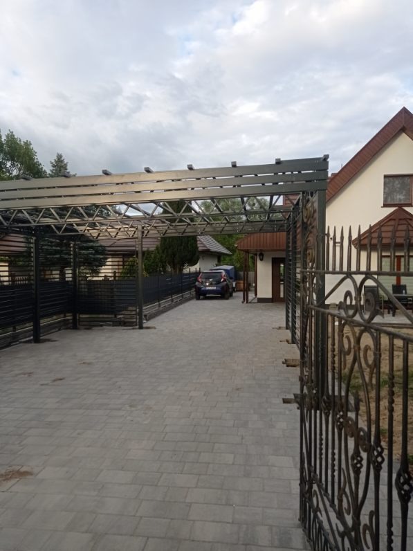 Metalowa konstrukcja zadaszenia nad podjazdem z widoczną bramą kutą i fragmentem domu w tle.