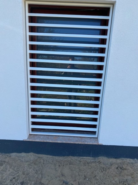 Biała, pozioma krata okienna na elewacji budynku, widoczna z zewnątrz, z odbiciem osoby w szybie.