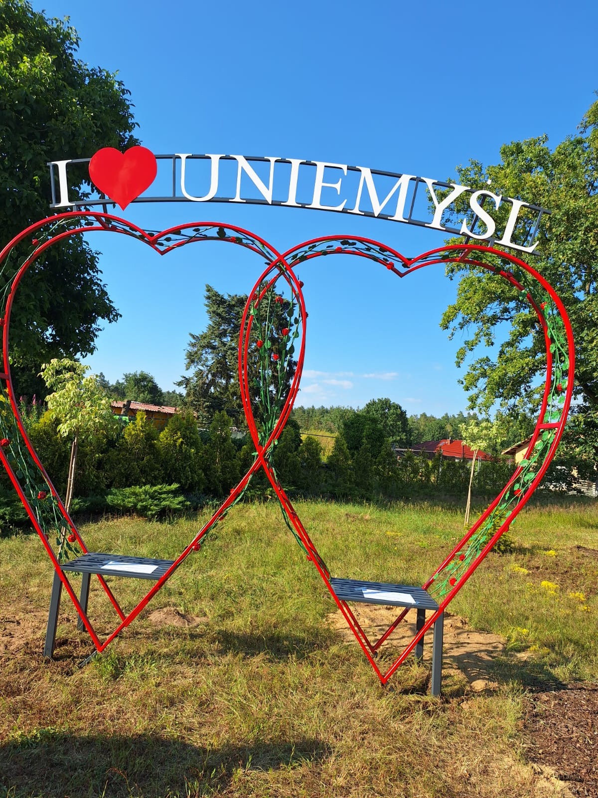 Dwa czerwone, metalowe serca tworzące bramę/pergole z miejscami do siedzenia, ozdobione zielonymi elementami imitującymi pnącza i czerwonymi kwiatami, z napisem 'I ❤️ UNIEMYŚL' u góry, na tle...