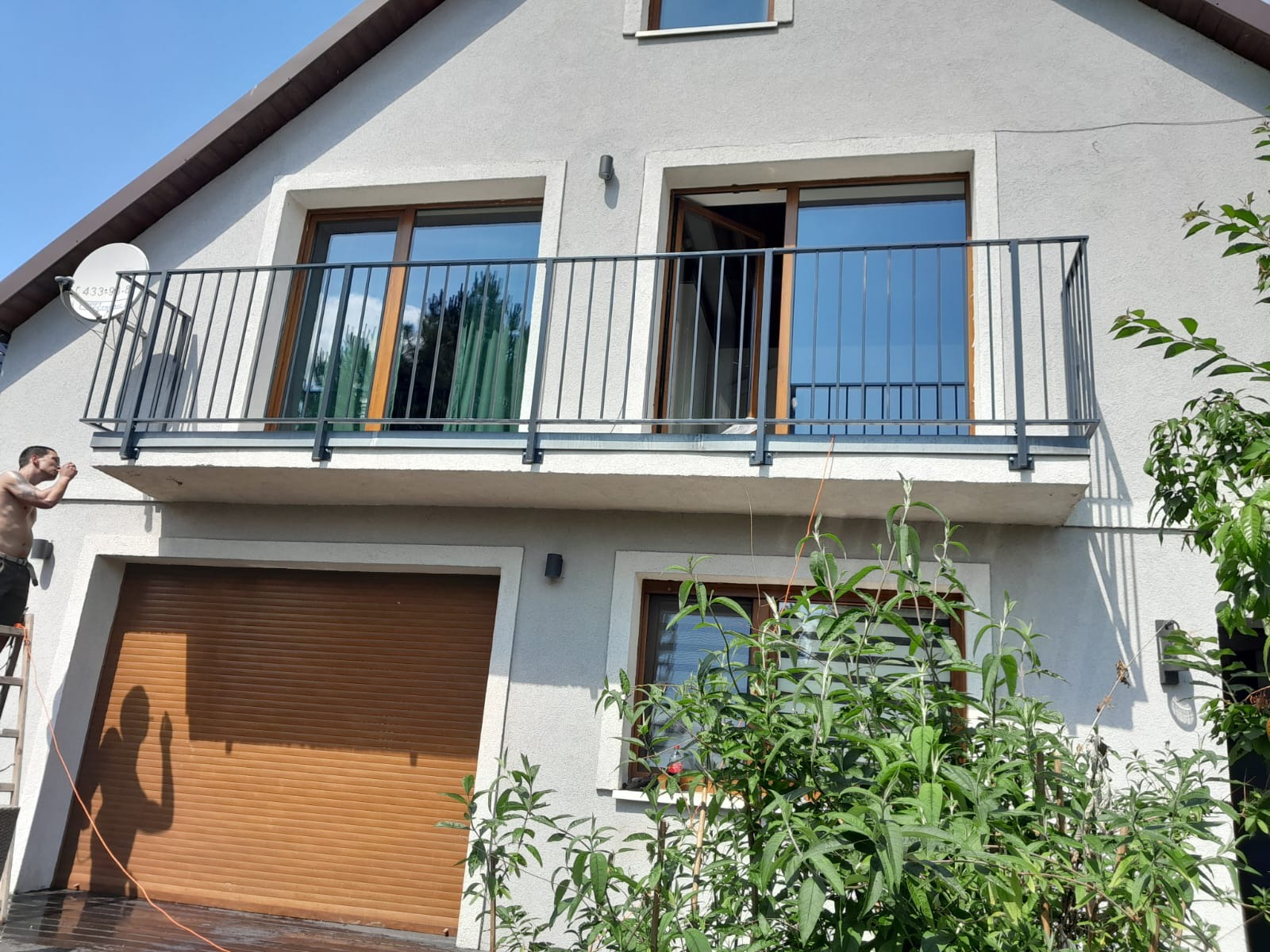 Szara elewacja domu z balkonem zabezpieczonym czarną, metalową konstrukcją; pracownik na drabinie maluje fragment elewacji