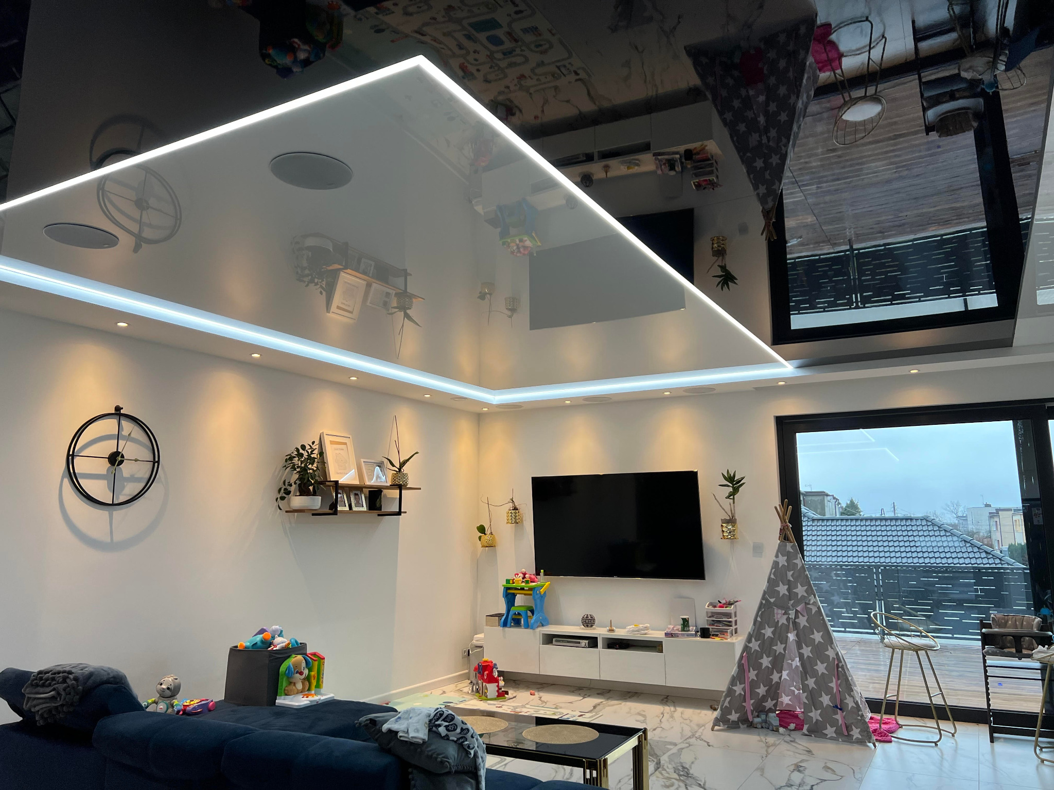 Nowoczesny salon z błyszczącym sufitem podwieszanym, oświetleniem LED i widocznymi głośnikami sufitowymi, odbijającym wystrój wnętrza.