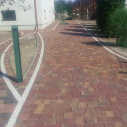 Nowo ułożona ścieżka z kostki brukowej w odcieniach czerwieni i brązu, obrzeża z białego grysu, prowadząca obok budynku z jasną elewacją i drewnianej stodoły.