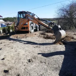 Pomarańczowa koparko-ładowarka CASE 580ST w trakcie pracy, łyżka zagłębiona w ziemi, świeże ślady wykopu, w tle drzewa i budynek w budowie.
