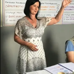 Kobieta w kwiecistej sukience prezentuje materiał szkoleniowy z hasłami dotyczącymi percepcji i emocji, widoczne notatki, długopisy i filiżanka kawy na stole.