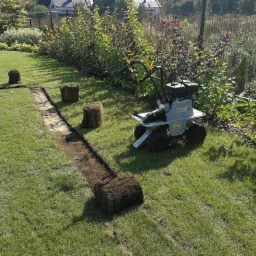Fragment trawnika z wykopanym rowkiem, obok leżą rolki trawy i stoi glebogryzarka, przygotowanie do ułożenia trawy z rolki.