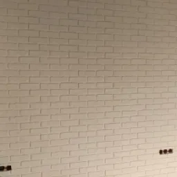 Ściana wykończona białą cegłą dekoracyjną, widoczne gniazdka elektryczne, fragment podłogi zabezpieczony kartonem.