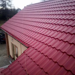 Czerwony dach z blachy falistej na jednopiętrowym domu, widok z góry, w tle zieleń i samochód z otwartym bagażnikiem.