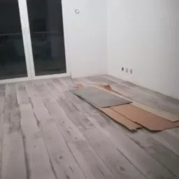 Jasne panele podłogowe ułożone w pokoju, z widocznymi fragmentami kartonów po panelach, leżącymi przy ścianie.