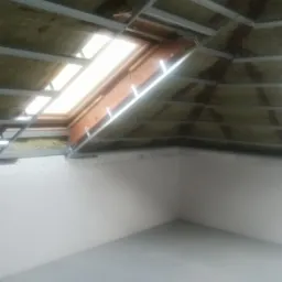 Poddasze w trakcie ocieplania z widoczną konstrukcją metalową, izolacją z wełny mineralnej i oknem dachowym, ściany pomalowane na biało, szara podłoga.