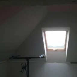 Poddasze z oknem dachowym w trakcie montażu płyt kartonowo-gipsowych, widoczny podnośnik do płyt, ślady różowej farby na ścianie.