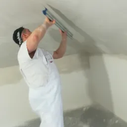 Pracownik w Pieckach w białym kombinezonie i czapce z daszkiem, wygładza sufit szpachlą, przygotowując go do malowania.