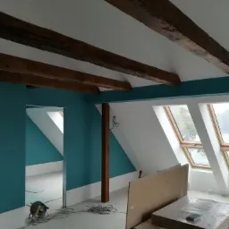 Poddasze w trakcie prac wykończeniowych: pomalowane na turkusowo ściany, drewniane belki stropowe, okna dachowe z widokiem na jezioro, widoczne narzędzia i materiały budowlane.