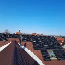 Panele słoneczne zamontowane na brązowym dachu domu jednorodzinnego z widocznym oknem dachowym i kominem, częściowo pokrytym śniegiem, na tle błękitnego nieba.