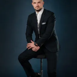 Portret mężczyzny w ciemnozielonym garniturze w kratę, białej koszuli i szarej kamizelce, siedzącego na stołku barowym na tle ciemnoniebieskiego tła. Na lewym nadgarstku widoczny zegarek z brązowym...