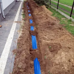 Wąski wykop wzdłuż chodnika z ułożonymi w regularnych odstępach niebieskimi rurami osłonowymi, widok z góry na brązową ziemię i fragment domu.