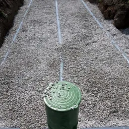 Drenaż rozsączający: widok na rury drenażowe ułożone w wykopie zasypanym żwirem, z zieloną studzienką kontrolną na pierwszym planie.