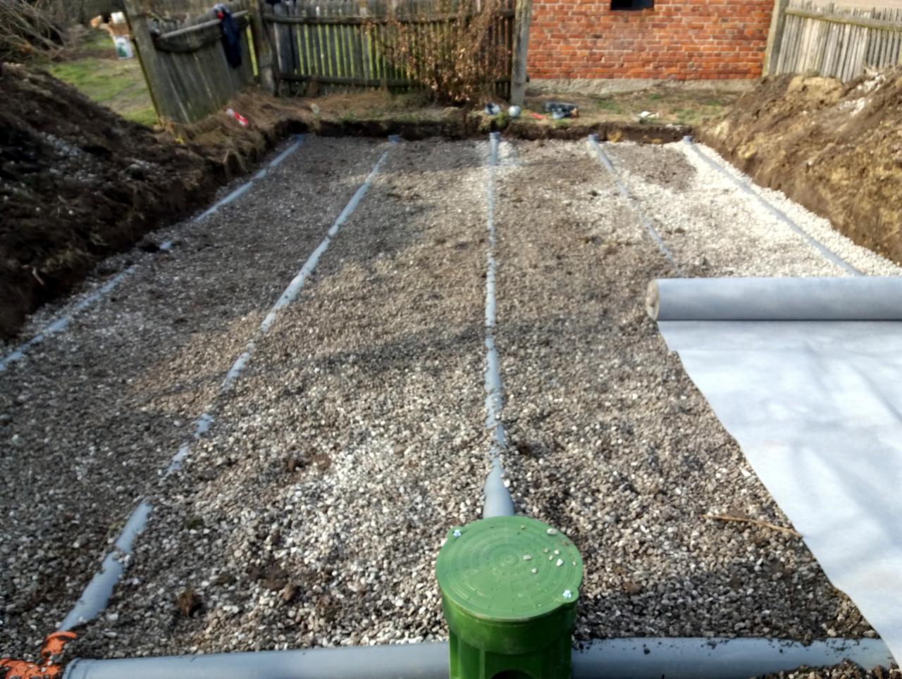 Instalacja rozsączająca ścieki w wykopie, rury drenażowe ułożone na żwirze, częściowo przykryte geowłókniną, widoczny zielony właz rewizyjny