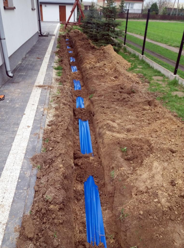 Wąski wykop wzdłuż chodnika z ułożonymi w regularnych odstępach niebieskimi rurami osłonowymi, widok z góry na brązową ziemię i fragment domu.