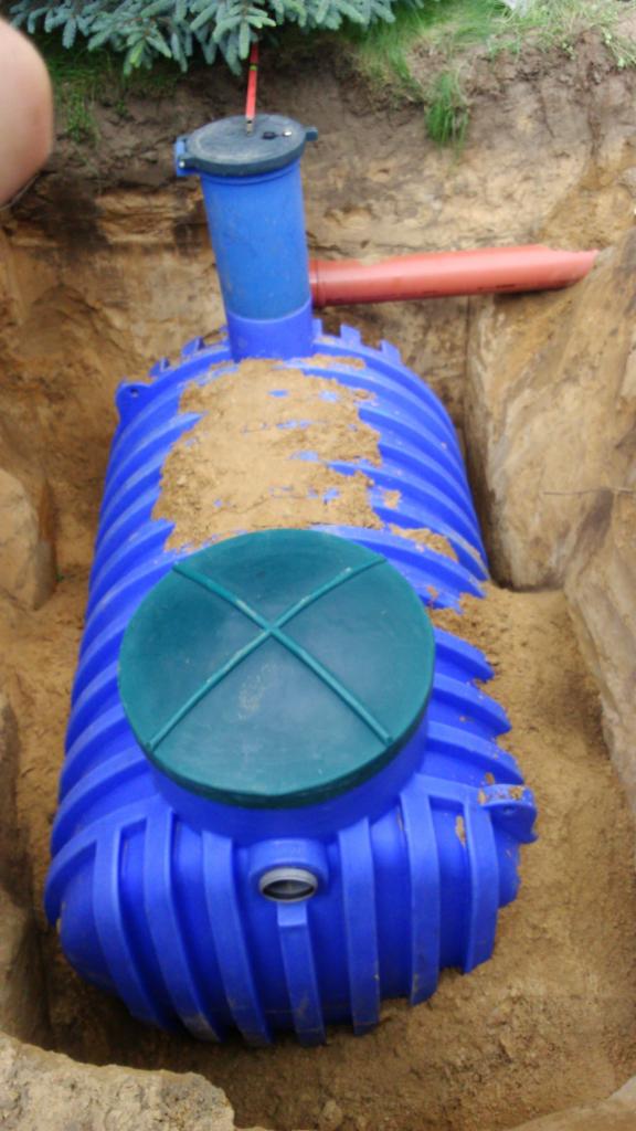 Niebieski zbiornik z zieloną pokrywą częściowo zasypany piaskiem w wykopie ziemnym, widoczna wystająca rura i fragment roślinności