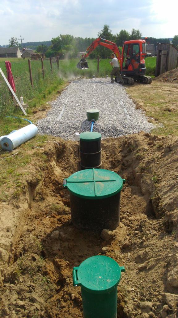 Instalacja przydomowej oczyszczalni ścieków w trakcie budowy: wykop z trzema zbiornikami z zielonymi pokrywami, rów wypełniony żwirem i pracujący mały koparka z operatorem.