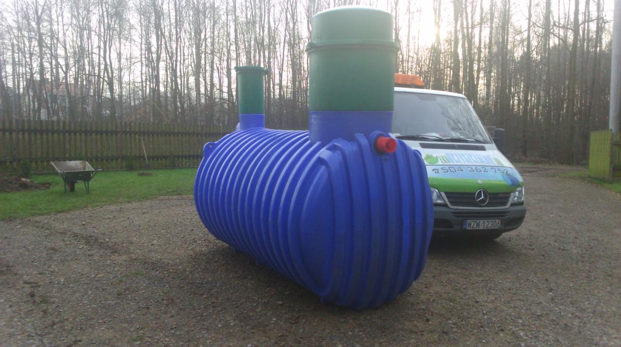 Gotowy, niebieski zbiornik na szambo z zielonymi pokrywami, ustawiony na żwirowym podłożu, na tle samochodu dostawczego firmy i zalesionego terenu.