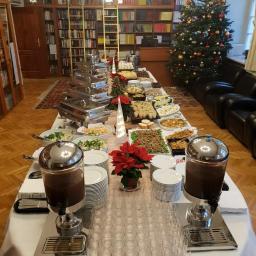 Catering Szafot - Ustawiony stół cateringowy z potrawami, napojami w dyspenserach, stosami talerzy i rzędami szklanek, w tle regały z książkami i choinka bożonarodzeniowa, dekoracje świąteczne na stole.