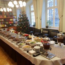 Catering Szafot - Stół cateringowy z podgrzewaczami, sałatkami i napojami w dyspenserach, ustawiony w pomieszczeniu z biblioteczką i choinką, przygotowany na firmową imprezę świąteczną.