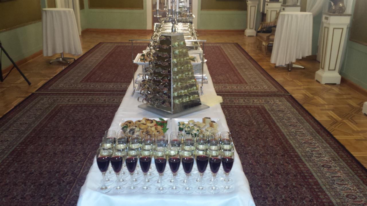 Długi stół z białym obrusem, na którym ustawione są rzędy kieliszków z czerwonym i białym winem, przekąski na talerzach oraz piramida z przekąskami, w tle parkiet i dywan.
