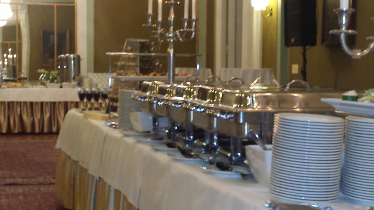 Bufet z podgrzewaczami, stosami talerzy i deserami w tle, ustawiony w eleganckiej sali z lustrami i żyrandolami.