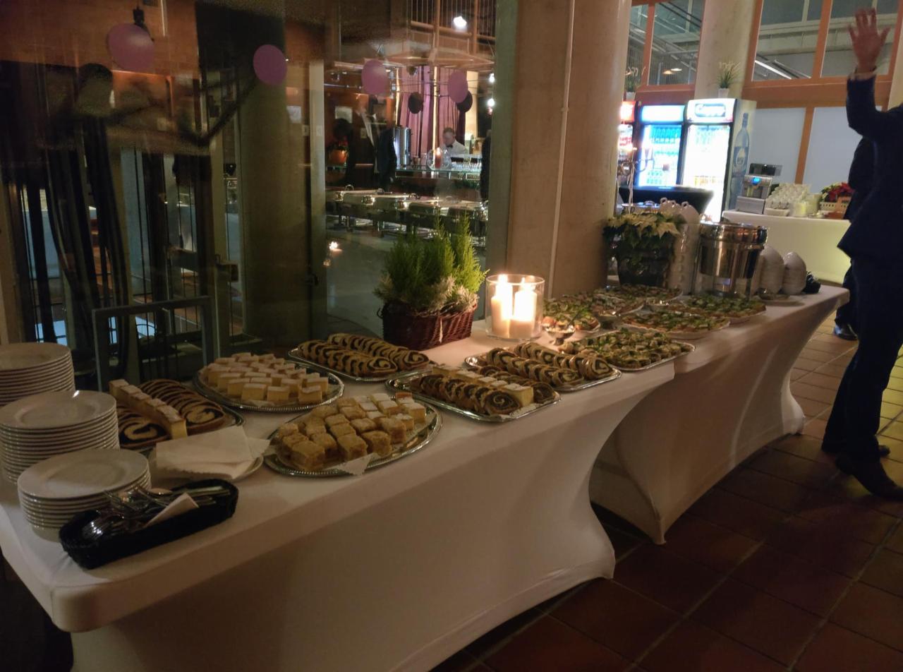 Stół cateringowy z ciastami, roladami makowymi i przekąskami, dekoracje roślinne i świece, w tle widoczni goście.