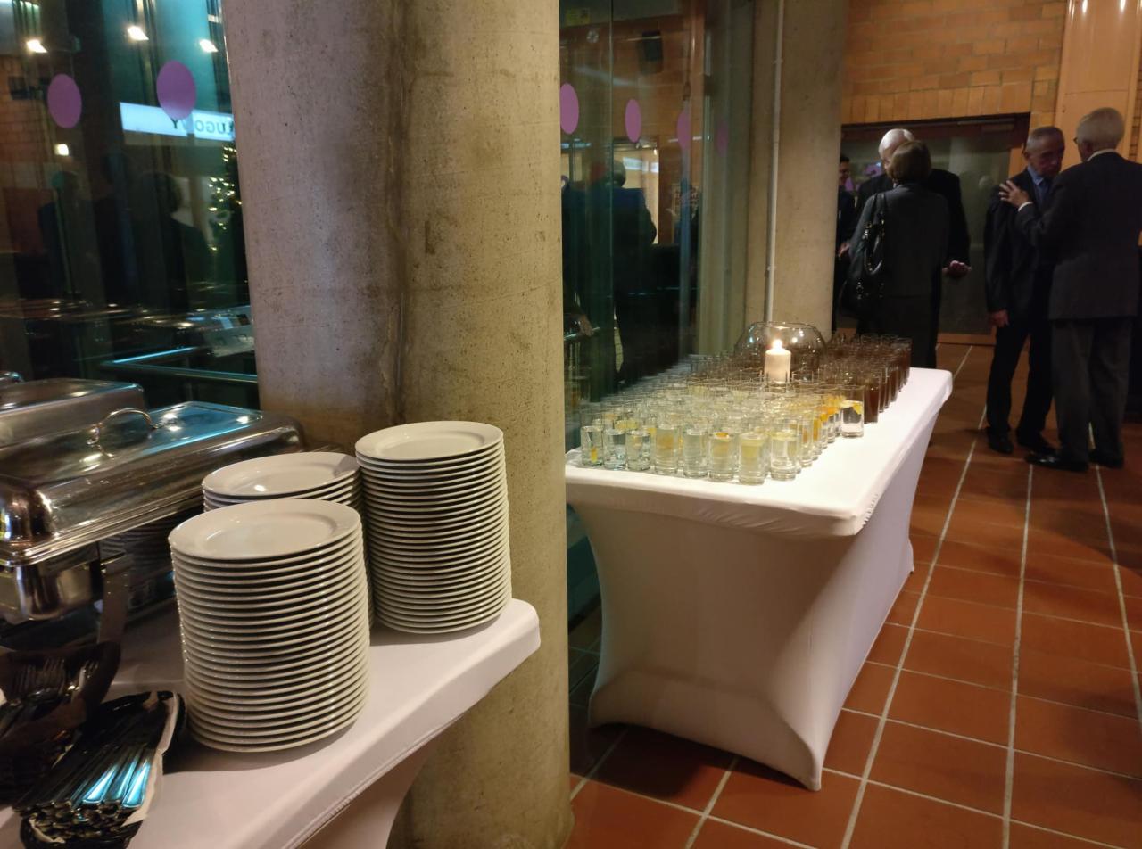 Przygotowany stół z ułożonymi szklankami z napojami, stosami talerzy i podgrzewaczem potraw, w tle rozmawiający goście w garniturach.