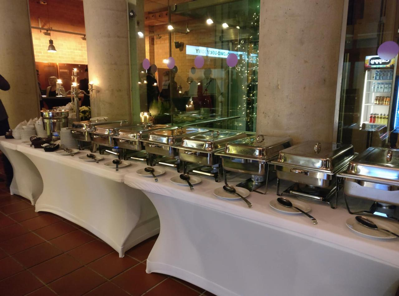 Stół szwedzki z podgrzewaczami i zastawą stołową, przygotowany do serwowania potraw podczas wydarzenia w Warszawie. W tle kolumny i dekoracje.