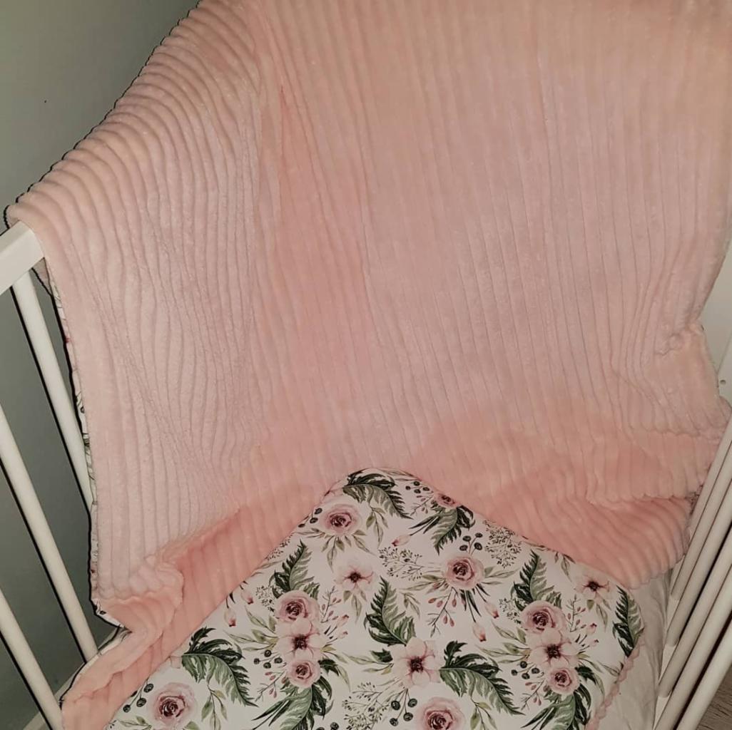Różowy, prążkowany kocyk i prześcieradło w kwiaty w białym łóżeczku dziecięcym.