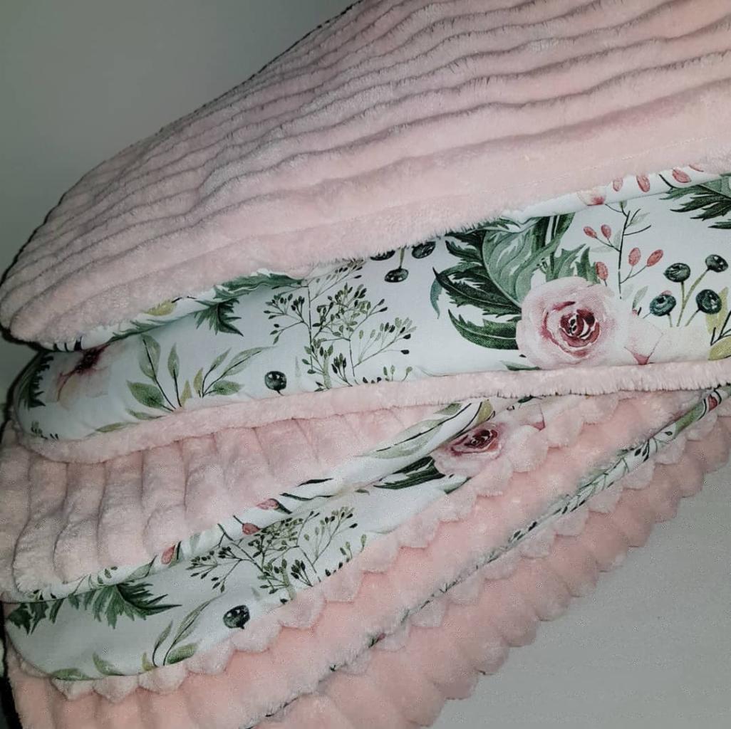 Stos różowych, pluszowych kocyków z bawełnianą lamówką w kwiaty, widoczne detale falbanki i faktury materiałów.