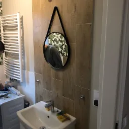 Łazienka z drewnopodobnymi płytkami, okrągłym lustrem na pasku, grzejnikiem drabinkowym, białą umywalką i widocznymi narzędziami oraz częścią osoby wykonującej prace.