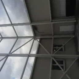 Wnętrze hali w trakcie budowy, widoczne białe stalowe elementy konstrukcyjne na tle szarej ściany z oknami i fragmentu sufitu, widok w górę na niebo z chmurami.