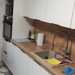 Biała zabudowa kuchenna z drewnianym blatem, zlewozmywakiem i baterią ze sprężyną; widoczne ściereczki i akcesoria kuchenne.