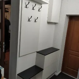 Biała zabudowa przedpokoju na wymiar z czarnymi akcentami: wieszak panelowy z półką, szafka wisząca i siedzisko ze schowkiem, widoczne drzwi wejściowe.