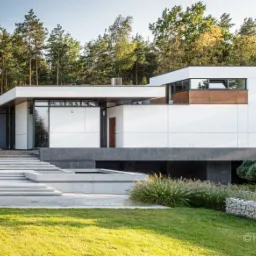 Nowoczesny dom z białymi elewacjami, dużymi przeszkleniami i schodami prowadzącymi do wejścia, otoczony zadbanym trawnikiem i zielenią, z widocznym gabionem z kamienia.