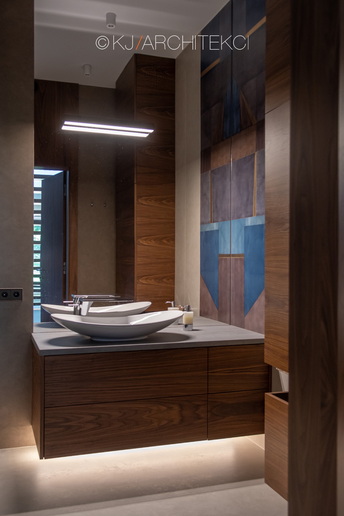 Nowoczesna łazienka z dwiema owalnymi umywalkami na blacie z szarego betonu, podświetlana szafka z ciemnego drewna, lustro z podłużną lampą i dekoracyjna ściana w geometryczne wzory.