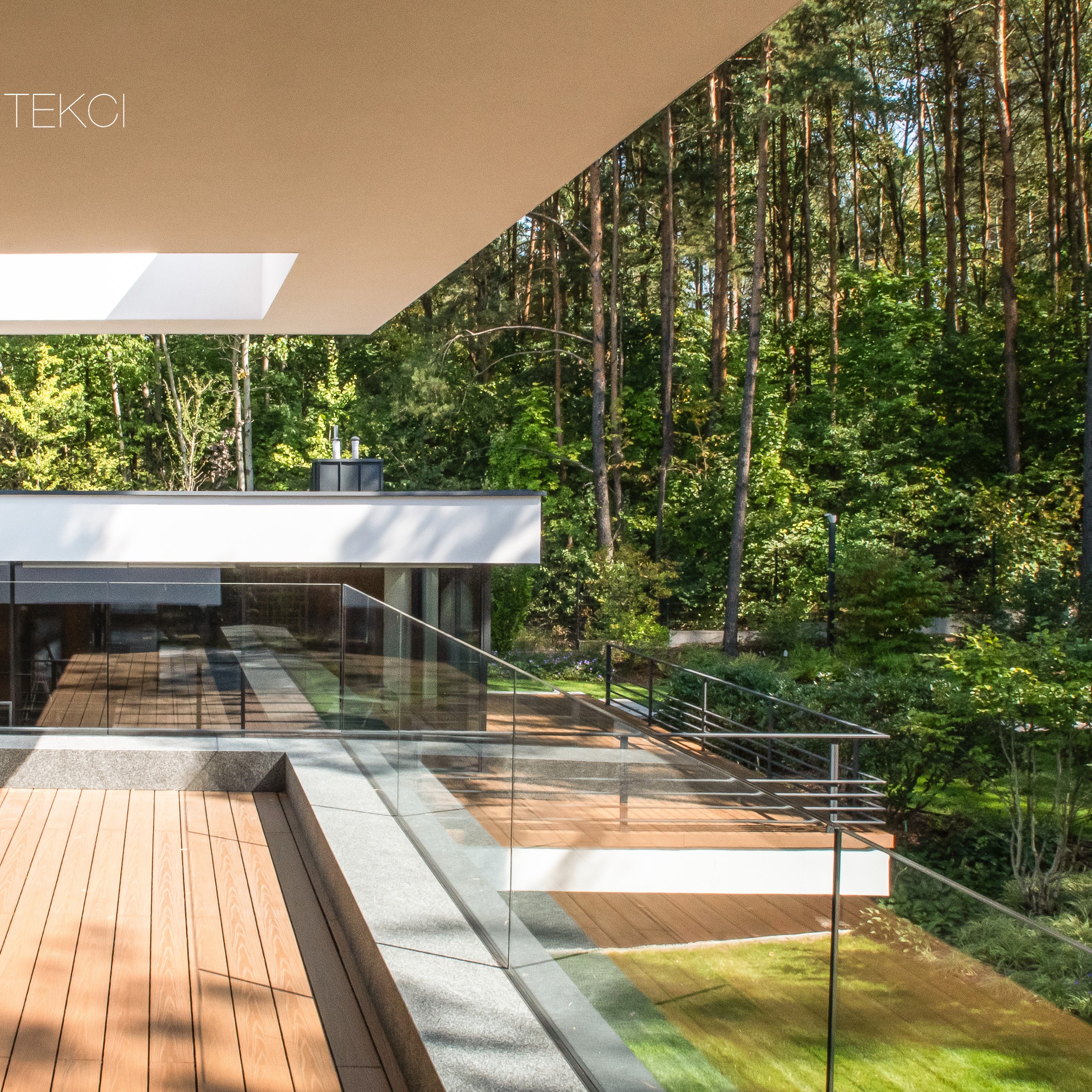 Nowoczesny dom z płaskim dachem i drewnianym tarasem, przeszklona balustrada, widok na las w tle.