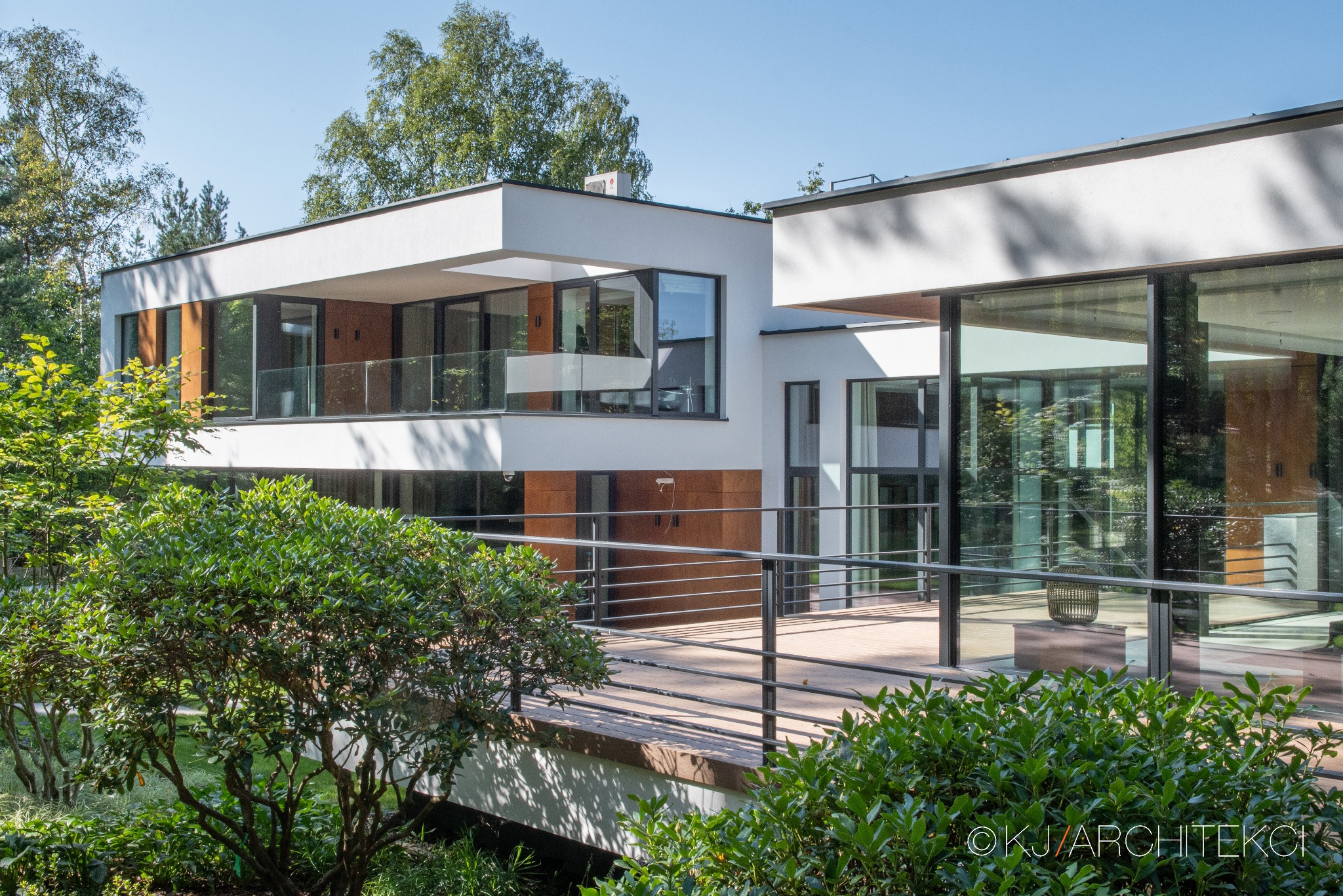 Nowoczesny, dwukondygnacyjny dom z płaskim dachem, dużymi przeszkleniami i drewnianymi panelami elewacyjnymi, otoczony zielenią.