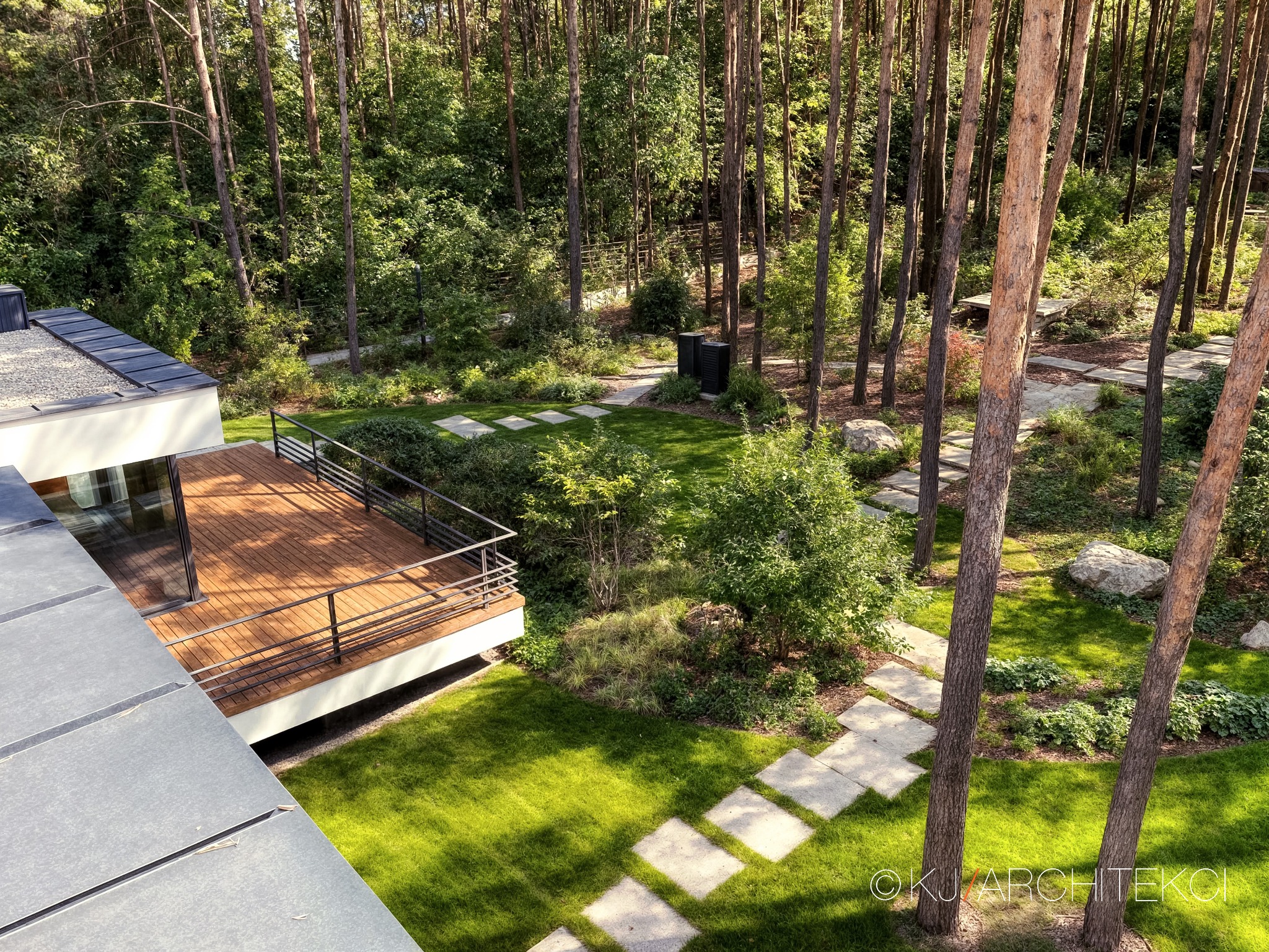 Nowoczesny dom z drewnianym tarasem i ścieżką z płyt betonowych prowadzącą przez zadbany trawnik w otoczeniu wysokich drzew iglastych.