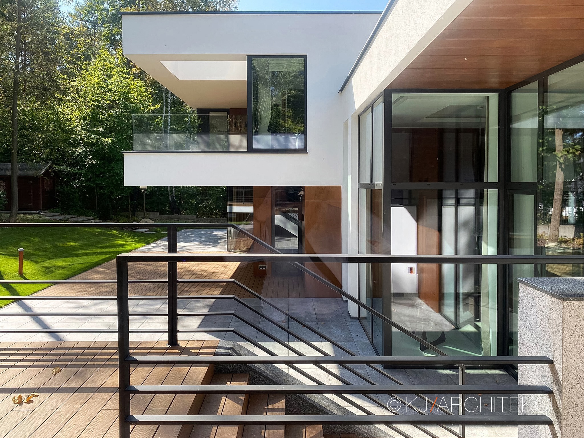 Nowoczesny dom z dużymi przeszkleniami, widok z drewnianego tarasu ze schodami, minimalistyczne balustrady, jasna elewacja i drewniane wykończenia.