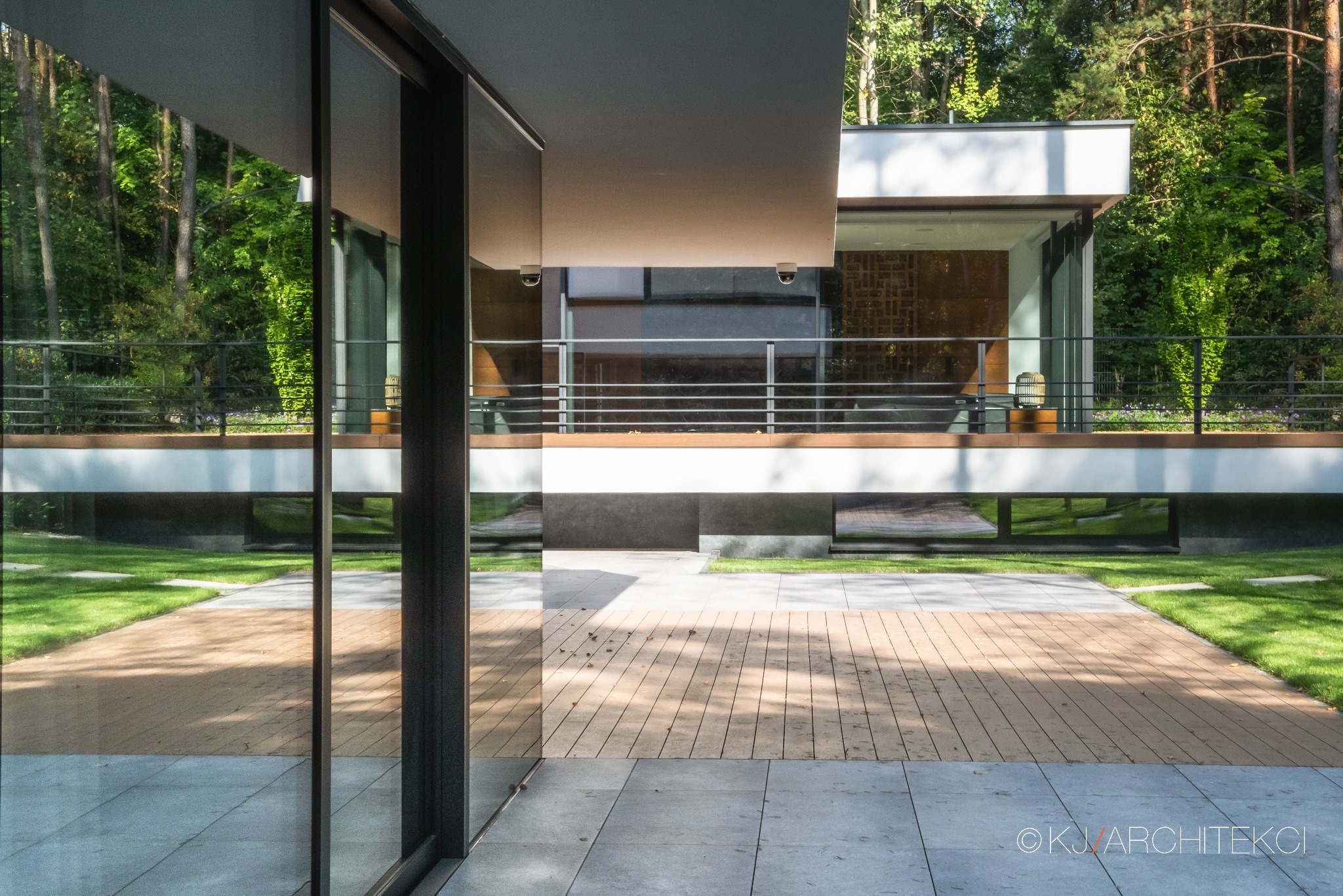 Nowoczesny budynek usługowy z dużymi przeszkleniami, widok z zewnątrz na taras z drewnianą podłogą i trawnik, otoczony drzewami.