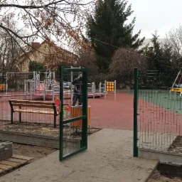 Nowo ogrodzony plac zabaw w Łajskach, z otwartą furtką, ławkami i częściowo rozpakowanymi urządzeniami zabawowymi, widoczne palety z kostką brukową.