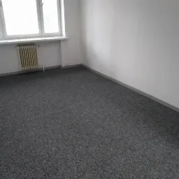 Pusty pokój z szarą wykładziną dywanową, białymi ścianami, grzejnikiem pod oknem i listwami przypodłogowymi. Widok z góry, oświetlenie dzienne.