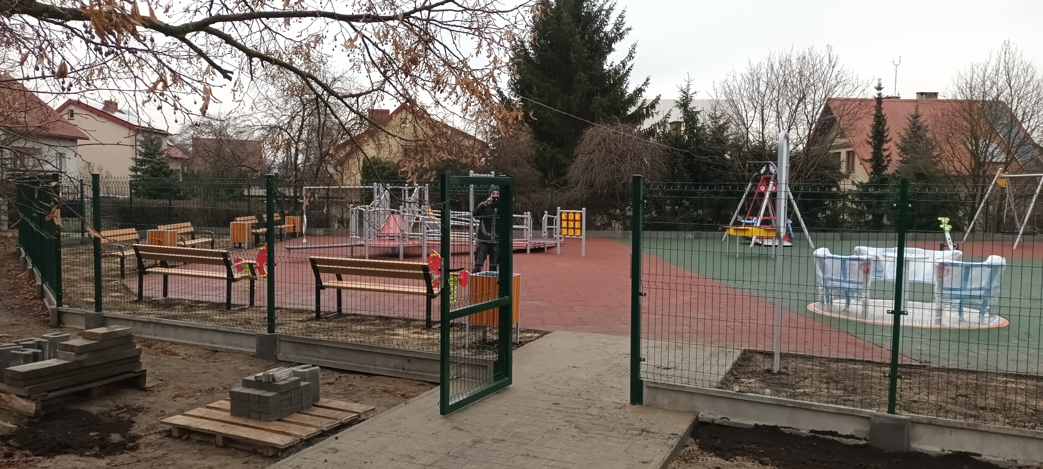 Nowo ogrodzony plac zabaw w Łajskach, z otwartą furtką, ławkami i częściowo rozpakowanymi urządzeniami zabawowymi, widoczne palety z kostką brukową.