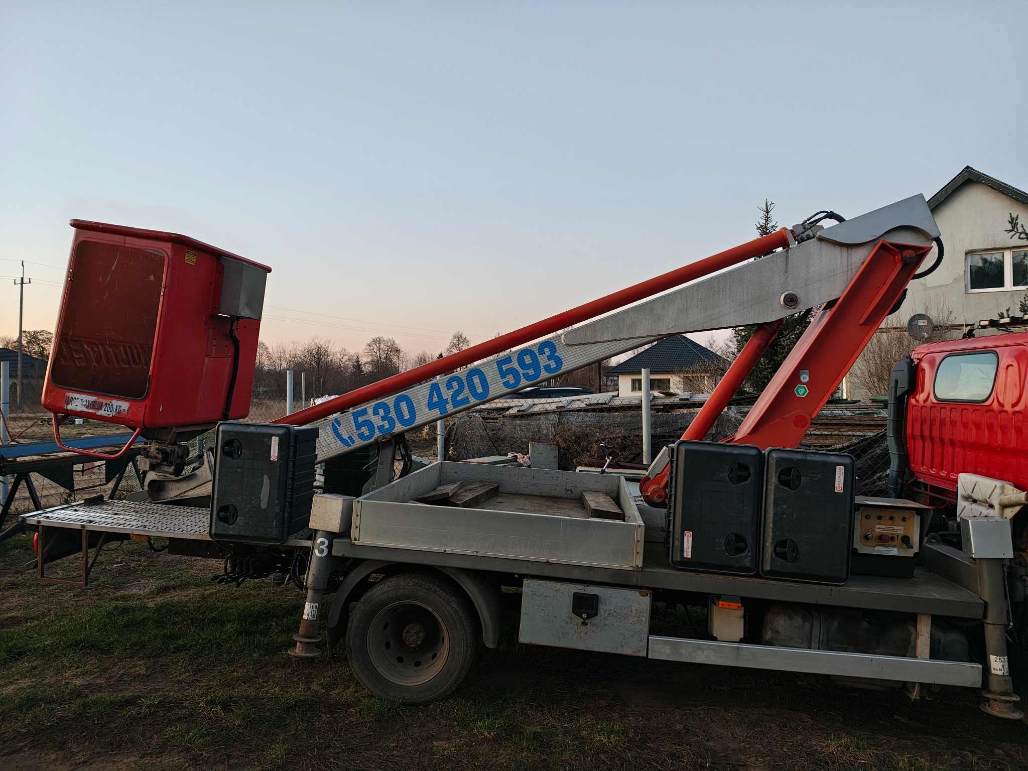 Czerwono-szara platforma robocza na podwoziu ciężarówki z numerem telefonu na ramieniu, zaparkowana na trawiastym terenie przed domem.