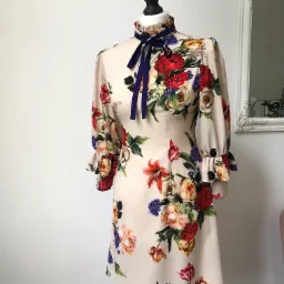 Sukienka z motywem kwiatowym na manekinie krawieckim, z granatową wstążką pod szyją, na tle białej ściany z lustrem w ozdobnej ramie.